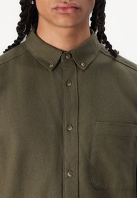 Camisa de color verde oliva con cuello abotonado, bolsillo en el pecho y tela texturizada que presenta patrones de tejido sutiles y botones marrones.