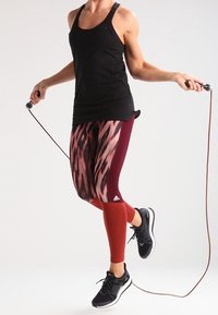 Top nero senza maniche, leggings a motivi bordeaux e rossi, e scarpe da ginnastica nere. Corda per saltare tenuta tra le mani con uno sfondo neutro.