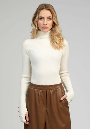 Pull à col roulé côtelé couleur ivoire, avec manches longues et ouvertures pour les pouces, associé à un pantalon en simili cuir marron avec ceinture élastique.