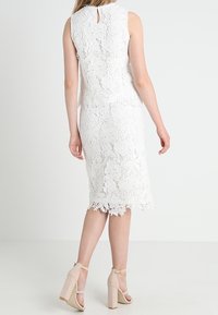 Vit spetsklänning med blommönster, knälång, ärmlös design. Har en nyckelhålsrygg och scalloped nederkant. Bärs med nakna klacksandaler.