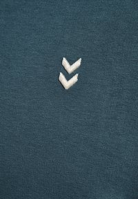 Camicia in cotone teal a maniche corte con tessuto strutturato e logo a zigzag ricamato di colore bianco sul lato sinistro.