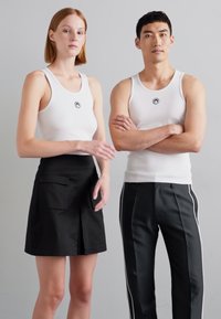 Biele rebrované tielka s kruhovým logom; model má na sebe čiernu sukňu s vreckami, ďalšia má čierne nohavice s bielymi bočnými pruhmi.