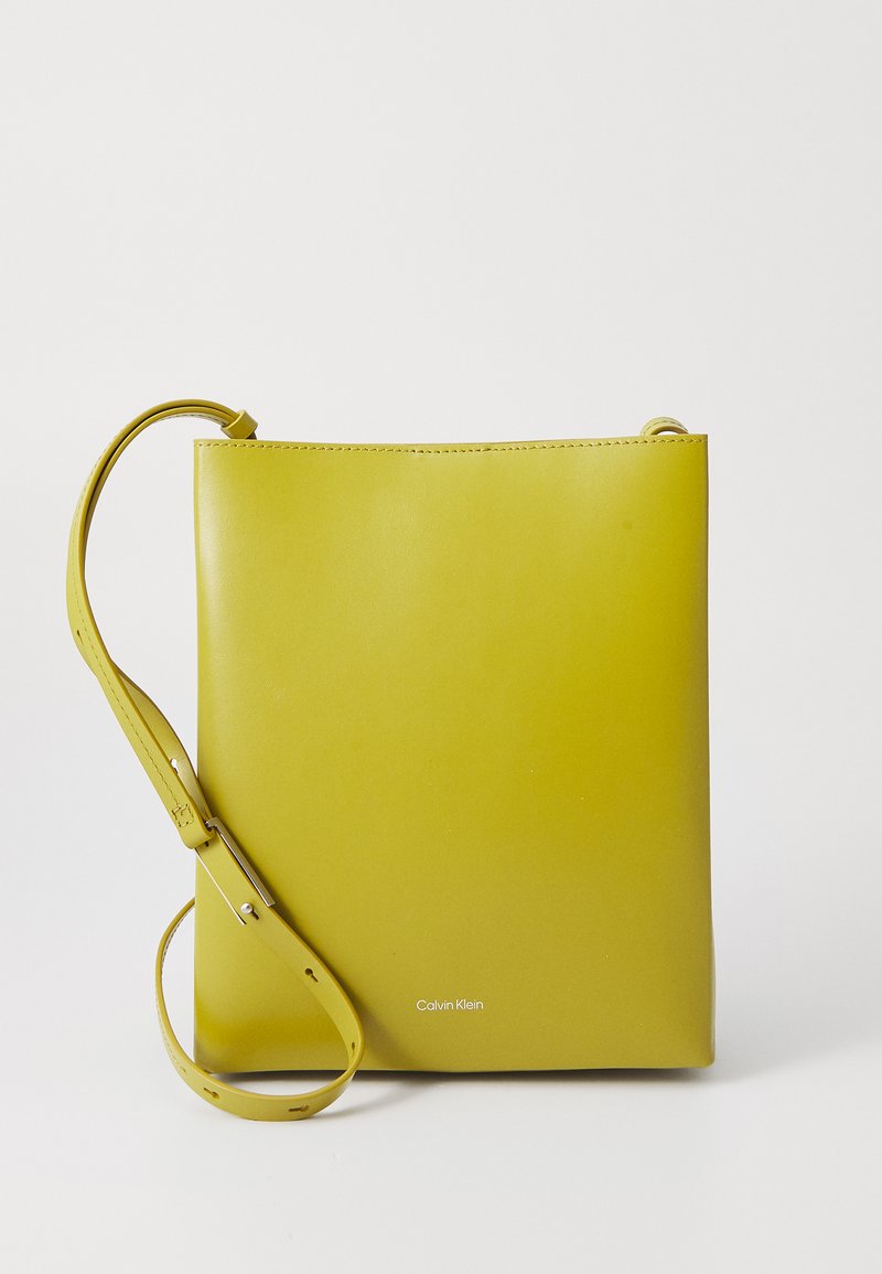 Žlutá kožená kabelka přes tělo s hladkým povrchem, minimalistickým designem a nastavitelným popruhem. Součástí je logo 'Calvin Klein' ve spodní části.