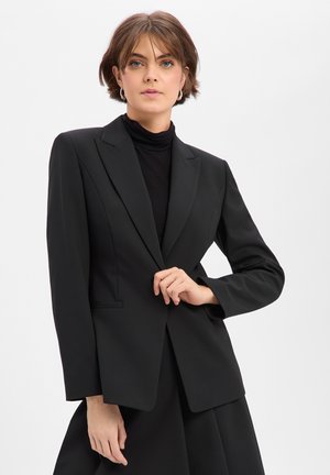 Jeune personne aux cheveux courts portant un blazer noir sur un col roulé noir, posant avec une main près du bouton de la veste.