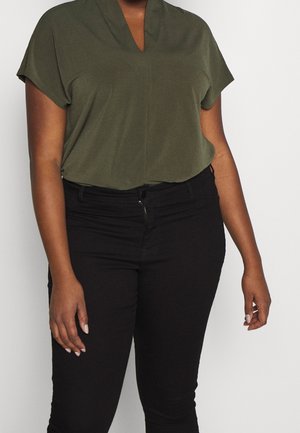 Oliwkowozielona bluzka z krótkim rękawem i dekoltem w serek, zestawiona z czarnymi dopasowanymi dżinsami. Materiał wydaje się miękki o gładkiej fakturze.