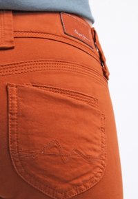 Pantalones de mezclilla naranja vistos de lado, con un bolsillo trasero que presenta un diseño de costura ondulada y un parche de cuero marrón en la cinturilla.