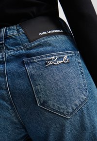 Džinsai su tamsiai mėlynu skalbimu, juodos odos etiketė su „KARL LAGERFELD“ logotipu ir sidabrinė „Karl“ segtukas ant užpakalinės kišenės.