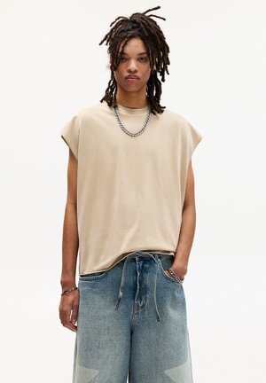 T-shirt beige sans manches avec une coupe décontractée, associé à un jean large bleu clair avec un motif délavé et une taille à cordon. Collier en chaîne argent.
