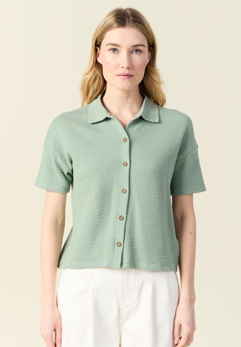 Camicetta a maniche corte con colletto in tessuto strutturato di un verde chiaro, con chiusura a bottone anteriore e tre bottoni marroni, abbinata a pantaloni bianchi.