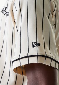 Men's short-sleeve jersey in cream with black vertical stripes, featuring a black embroidered logo on the sleeve and a textured finish.