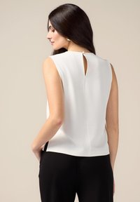 Blusa bianca senza maniche con un dettaglio a chiave sul retro, texture liscia e linee pulite. Abbinata a pantaloni neri, creando un look a contrasto.