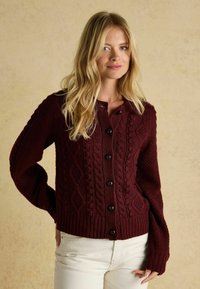 Burgundský kardigán s kabelkovým vzorom, s rebrovanými manžetami a lemom, obsahujúci šesť tmavých gombíkov vpredu. Nosený cez biele nohavice.