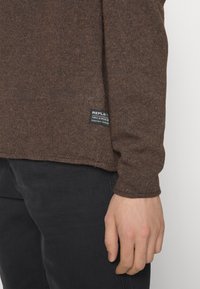 Gros plan d'une personne portant un pull en maille marron avec une petite étiquette noire près de l'ourlet, associé à un pantalon noir.