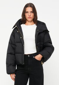 Zwarte pufferjack met capuchon, met een boxy vorm, gouden rits en drukknoopjes. Glad textuur met zichtbare horizontale stiksels.