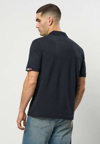 Mann med kort hår sett bakfra, iført en svart polotrøye med et lite italiensk flagg på venstre erme og lyseblå jeans.