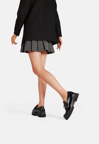 Scarpe in vernice nera con suola spessa e dettaglio a fiocco decorativo, indossate con una gonna grigia a pieghe e un blazer nero.