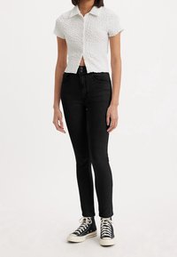 Person som bär en texturerad vit kortärmad skjorta, högmidjade svarta tightjeans och svarta höga sneakers med vita snören.