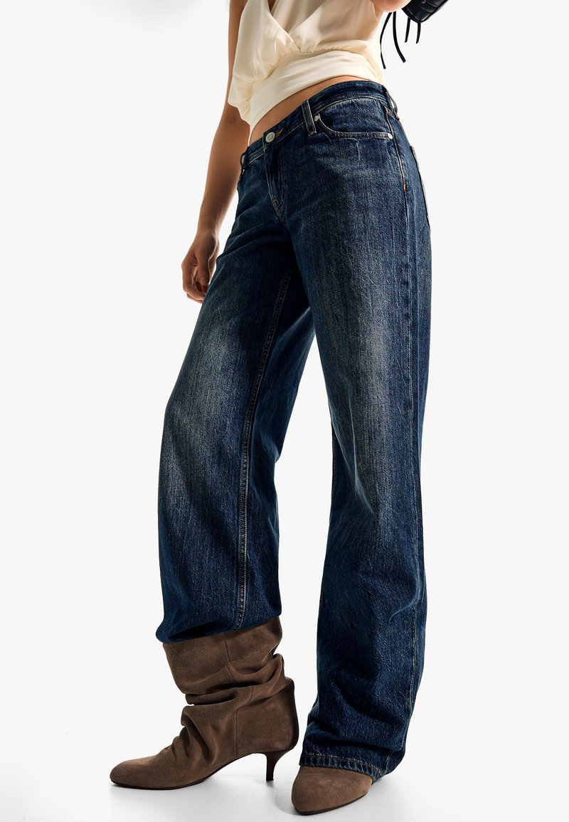 Nő magas derekú, bőszárú, sötétkék farmert és barna, magassarkú velúr csizmát visel, világos színű rövid felsővel.