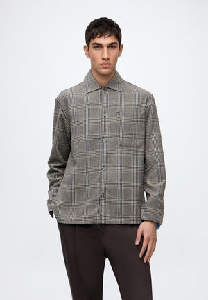 Jeune homme portant une chemise à carreaux marron et beige avec col et boutons, ainsi qu’un pantalon marron foncé, debout devant un fond clair uni.