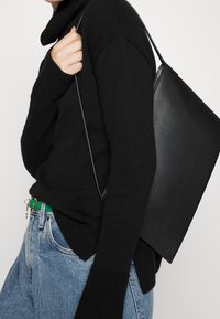 Black leather tote bag with a minimalist design, featuring a smooth texture and long shoulder strap, paired with a black knitted sweater.