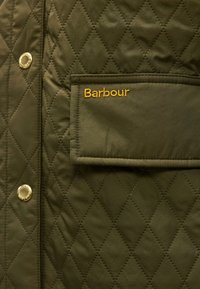 Πράσινο μπουφάν με καπιτονέ ύφασμα σε σχέδιο ρόμ βου, χρυσά κουμπιά με πατήματα και ορθογώνια θήκη με καπάκι κεντημένο με τη λέξη "Barbour" σε κίτρινο χρώμα.