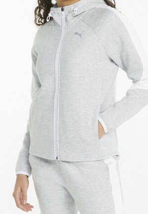 Persoon draagt een lichtgrijze hoodie met rits en witte strepen op de mouwen en bijpassende broek, hand in de zak, gecropt vanaf de nek tot halverwege het bovenbeen.
