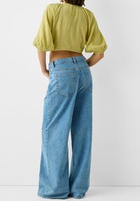 Top corto verde con maniche a sbuffo abbinato a jeans a gamba larga azzurri chiari con tasche posteriori e una texture leggermente sbiadita.