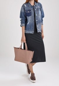 Kvinna som bär en sliten denimjacka över en svart midiklänning, bär en stor beige axelväska och har på sig bruna knutna tofflor.