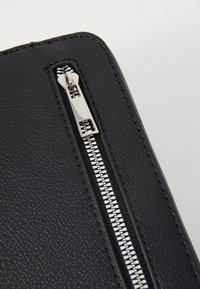 Étui en cuir noir texturé avec une fermeture éclair argentée et une tirette lisse, mettant en avant un design minimaliste et un savoir-faire de qualité.