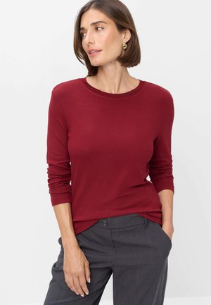 Frau mit kurzen braunen Haaren, die ein rotes langärmeliges Oberteil und graue Hose trägt, steht mit einer Hand in der Tasche vor einem weißen Hintergrund.