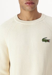 Cremefarbener Rippstrickpullover mit rundem Ausschnitt, Raglanärmeln und einem grünen Krokodil-Logo auf der linken Brust. Weicher Griff.