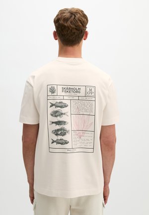 Homme portant un t-shirt beige avec un graphisme noir et rose d’illustrations de poissons et un texte dans le dos, debout sur un fond blanc.