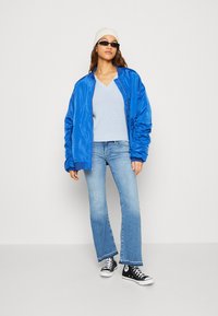 Blå sateng bomberjakke, lys blå strikket topp, lys blå vide jeans, svarte kanvas sneakers, hvit lue og solbriller.