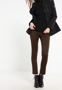 Veste noire matelassée avec des zippers, associée à un haut noir à manches longues et un pantalon moulant marron, complétée par des bottines noires.