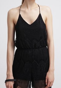 Femme portant un combishort sans manches en dentelle noire avec des bretelles spaghetti et une taille cintrée, se tenant devant un arrière-plan clair.