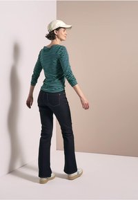 Gestreiftes grünes Langarmoberteil, schwarze ausgestellte Jeans, weiße Sneakers. Abgerundet mit einer hellen Mütze. Modell, das von hinten vor einem neutralen Hintergrund steht.