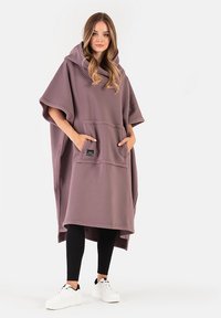 Poncho w dużym rozmiarze w stonowanym kolorze mauve, wykonane z miękkiego materiału. Posiada dużą przednią kieszeń, szeroki kaptur i krótkie rękawy. Noszone z legginsami i sportowymi butami.