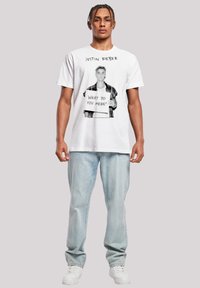 Weißes Baumwoll-T-Shirt mit einem schwarz-weißen Grafikdruck einer Person, die ein Schild mit dem Text "Was meinst du damit?" hält. Lässiger Schnitt.