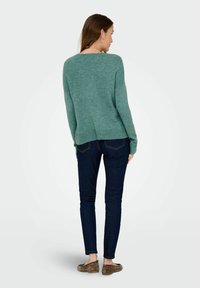Donna di spalle che indossa un maglione verde, jeans skinny blu scuro e mocassini in suede marroni, su uno sfondo chiaro e semplice.