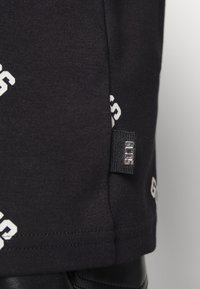 Camiseta de algodón negra con patrón de logo blanco; presenta una etiqueta en el dobladillo con el nombre de la marca. Textura suave, mangas cortas y un corte informal.