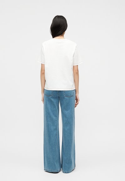 Γυναίκα με μακριά σκούρα μαλλιά που στέκεται, φορώντας ένα απλό άσπρο t-shirt με κοντά μανίκια και μπλε τζιν παντελόνι ψηλόμεσο με φαρδιά μπατζάκια, μπροστά σε λευκό φόντο.