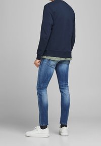 Mörkblå sweatshirt med rund halsringning, lager på en olivgrön skjorta, matchad med åtsittande blå jeans och vita sneakers.