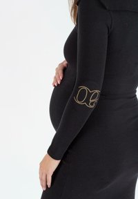 Schwarzes geripptes Kleid mit langen Ärmeln, versehen mit einem goldenen Logo am Ellbogen. Enganliegendes Design betont die Silhouette. Weicher Stoff.