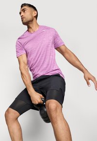 Športový outfit pozostávajúci z ľahkého, krátkeho ružového trička s textúrovaným vzorom a čiernych šortiek s sieťovinovými akcentmi. Drží kettlebell.