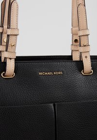 Mustapahuisena käsilaukku, jossa on teksturoitu pinta, beige olkahihnoja ja kultaiset yksityiskohdat. Logo esillä etupuolella. Siisti, strukturoitu muotoilu.