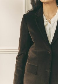 Barna kordbársony blézer karcsúsított szabással, bevágott hajtókákkal, gombos záródással és elülső zsebbel, fehér csipkés blúz fölött viselve.