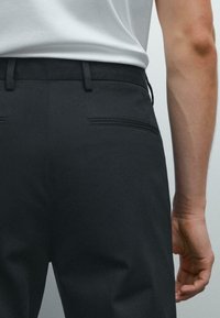 Persoon in een zwart op maat gemaakt broek en een wit shirt, gezien vanaf de onderrug met een arm zichtbaar naast het lichaam.