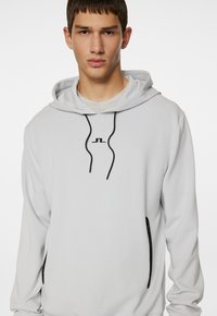 Lys grå pullover hoodie med snor i huen, prydet med et sort logo i midten og sorte lynlåslommer på siden. Blød stoftekstur.
