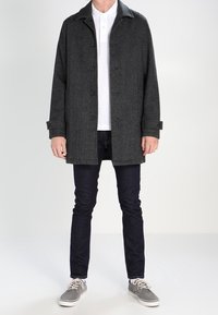 Manteau en laine gris avec un col cranté, des boutons frontaux et des poignets boutonnés, porté par-dessus un polo blanc et un jean sombre avec des baskets grises.