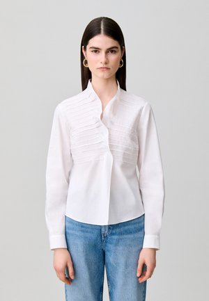 Femme aux cheveux lisses et foncés portant un chemisier blanc plissé et un jean bleu, debout face à l’avant sur un fond gris clair uni.